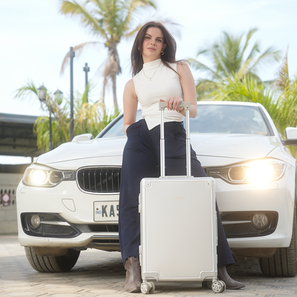 A woman standing beside a white full aluminium trolley cabin luggage with a car in the background.